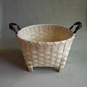 May include: A round woven basket made of light-colored wood strips featuring a reinforced rim, four short wooden feet at the base, and two dark brown leather handles attached with metal rivets on opposite sides of the upper edge.