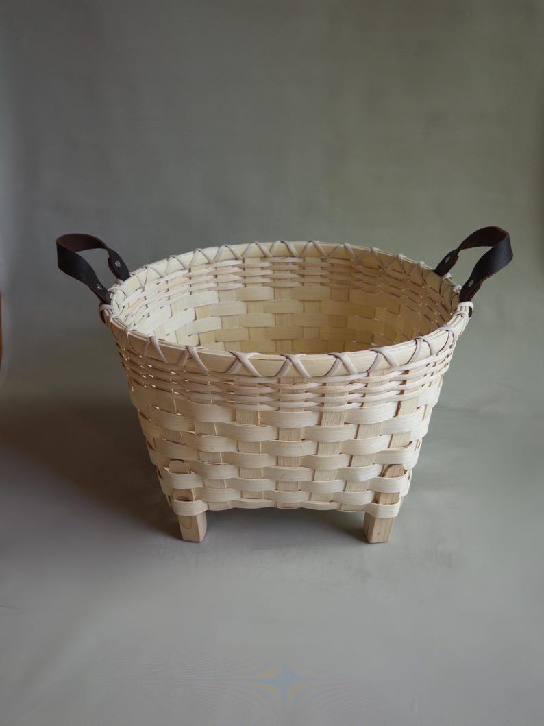 May include: A round woven basket made of light-colored wood strips featuring a reinforced rim, four short wooden feet at the base, and two dark brown leather handles attached with metal rivets on opposite sides of the upper edge.