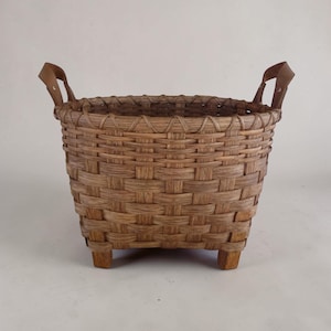 May include: A square, woven basket with a natural brown finish. The basket has a rectangular shape, raised on four small wooden feet. It features two leather handles and is suitable for storage or display.