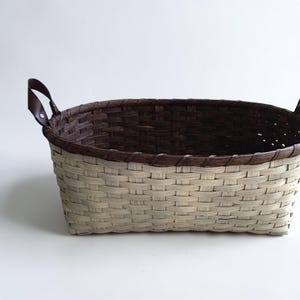 May include: Woven basket with a rectangular shape. The basket features a two-tone design, with a light beige exterior and a dark brown interior and rim. It has two brown leather handles.
