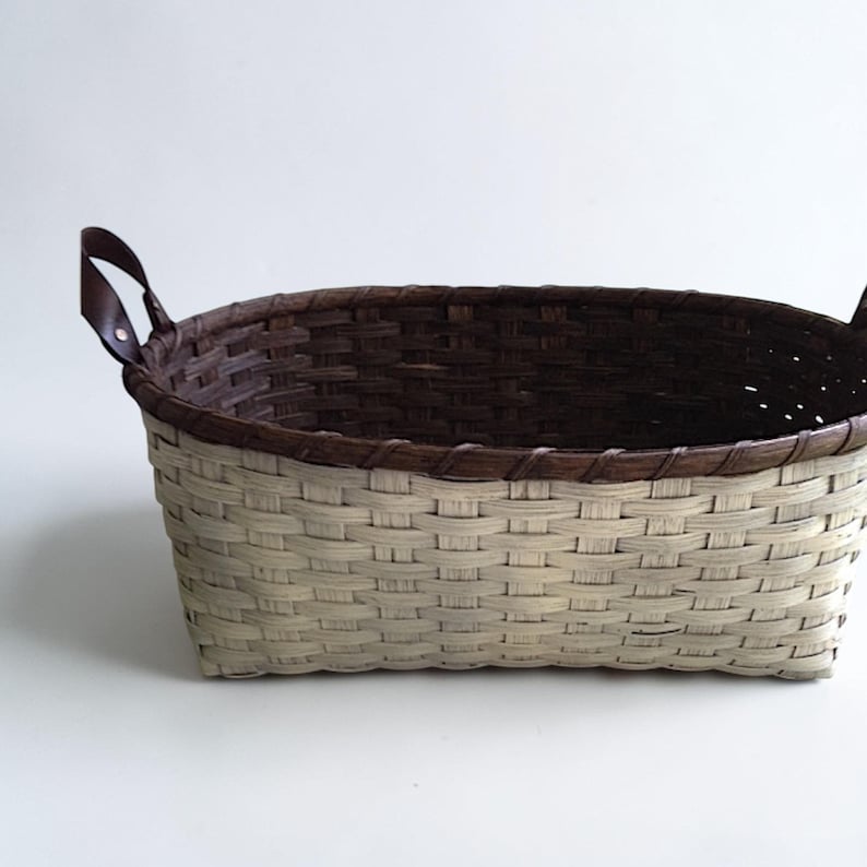 May include: Woven basket with a rectangular shape. The basket features a two-tone design, with a light beige exterior and a dark brown interior and rim. It has two brown leather handles.