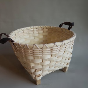 May include: A woven light-colored wood basket with a square base and rounded top rim. The basket features a cross-weave pattern, four small wooden feet at the base, and two dark brown leather handles attached to the sides with metal rivets.