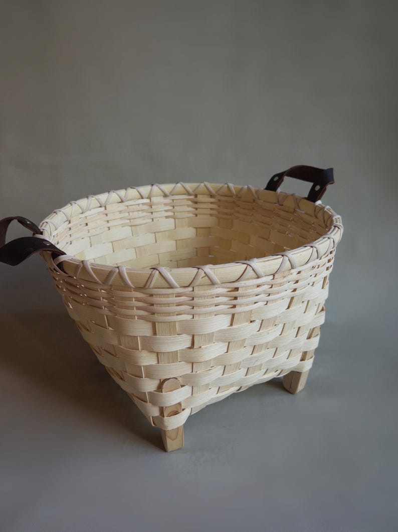 May include: A woven light-colored wood basket with a square base and rounded top rim. The basket features a cross-weave pattern, four small wooden feet at the base, and two dark brown leather handles attached to the sides with metal rivets.