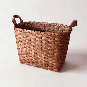 May include: A rectangular brown woven basket with two leather handles. The basket has a textured surface and a decorative woven trim around the top edge. The basket is empty and sits on a white surface.