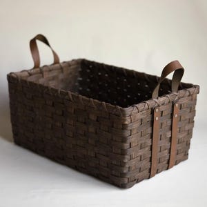 May include: A rectangular woven basket in a dark brown finish featuring a flat base and vertical sides. The basket includes two brown leather handles attached to the exterior with silver-toned metal rivets on each side for secure handling.