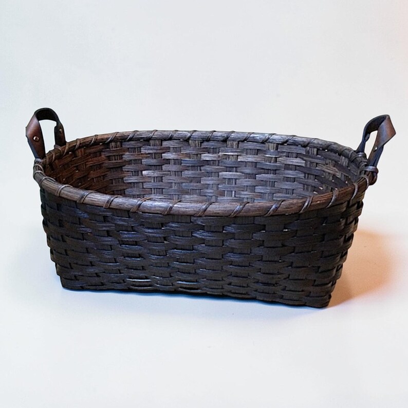 May include: A dark brown woven basket with a rectangular shape and leather handles. The basket is made of tightly woven strips of material, creating a textured surface. The handles are attached to the sides with metal rivets.