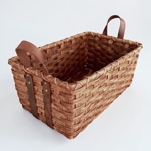 May include: A rectangular woven basket with brown leather handles and straps. The basket is made of light brown woven material and has two arched handles. The leather straps are attached with brass-colored rivets.