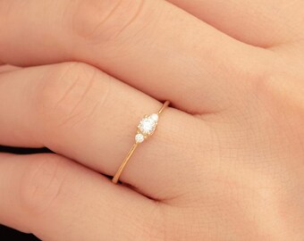 May include: A delicate gold ring featuring three small, sparkling gemstones. The ring is thin and elegant, with the gemstones clustered together in a subtle design. The ring is shown on a person's finger, highlighting its fine details and minimalist style.