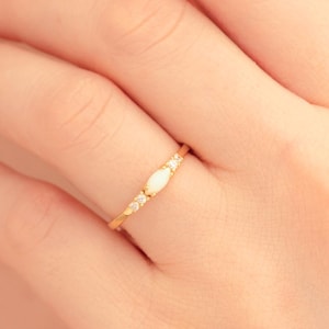 May include: A gold ring featuring a central oval-shaped opal gemstone, flanked by smaller, round, clear gemstones. The ring is set on a delicate gold band, creating a minimalist and elegant design. The ring is shown on a person's finger.