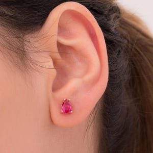 May include: A close-up of a person's ear with a small, teardrop-shaped, ruby-colored stud earring. The earring is set in a gold-colored metal. The background is a soft, neutral color.