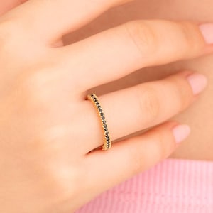 May include: A gold ring with a band of small, black gemstones. The ring is on a finger, with the hand and arm in the frame. The ring is a simple, elegant design, perfect for everyday wear or special occasions.