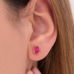 May include: A close-up of a gold stud earring with a teardrop-shaped, bright pink gemstone. The earring is set in a four-prong setting. The earring is on a person's ear.