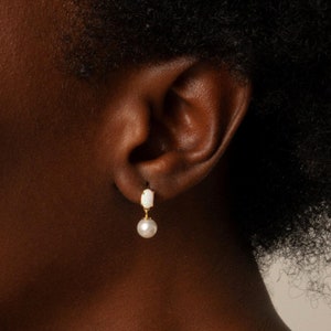 May include: Gold-tone earrings with a white, elongated top and a dangling pearl. The earrings are worn on a person's ear, showcasing the jewelry's design and elegance. The earrings are a classic accessory.