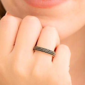 May include: A gold-toned ring with three rows of small, black gemstones. The ring is worn on a finger, showcasing its design and the contrast between the gold and black elements. The ring's design is simple yet elegant.