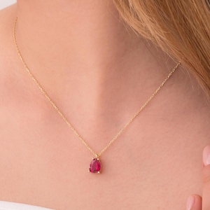 May include: A delicate gold necklace featuring a teardrop-shaped, vibrant red gemstone pendant. The pendant is suspended from a fine gold chain, creating a simple yet elegant design. The necklace is worn against a white top.