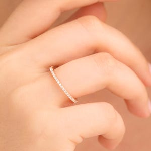 May include: A delicate gold ring with a thin band, adorned with a continuous row of small, sparkling diamonds. The ring is shown on a person's finger, highlighting its elegant and minimalist design. This piece is perfect for everyday wear or special occasions.
