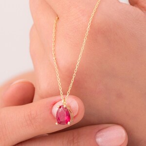 May include: A gold necklace featuring a teardrop-shaped, red gemstone pendant. The pendant is held by four prongs. The necklace has a delicate chain. The background is a soft, neutral colour.