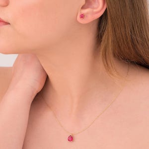 May include: A gold necklace featuring a teardrop-shaped red gemstone pendant. Matching gold stud earrings with red gemstones are also visible. The jewellery is set against a neutral background.