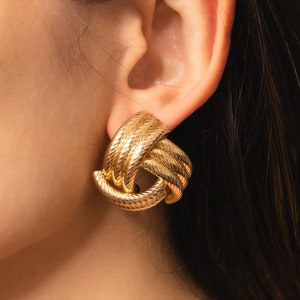May include: Gold knot-shaped earrings. The earrings are made of a braided metal and have a shiny finish.
