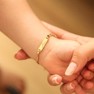 May include: A gold-colored bracelet with the name "Alice" engraved on a rectangular plate. The bracelet is on a child's wrist, held by an adult's hand. The bracelet has a curb chain design.
