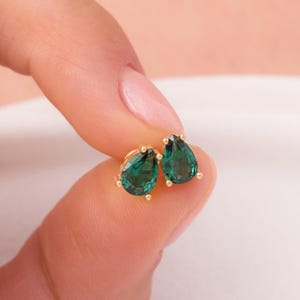 May include: A pair of gold-colored stud earrings, each featuring a teardrop-shaped, emerald-green gemstone. The earrings are held between fingers, showcasing their polished finish and vibrant color. The setting appears to be a classic four-prong design.