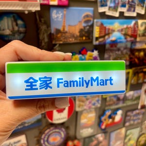 May include: A rectangular magnet with the text "全家 FamilyMart" in blue and white. The top of the magnet is green. The magnet is held in front of a collection of other magnets on a refrigerator.