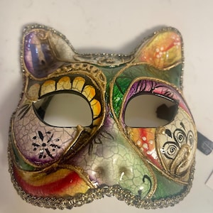 Pode incluir: Uma máscara colorida em forma de gato com acabamento dourado e detalhes brilhantes. A máscara apresenta uma variedade de cores, incluindo verde, roxo, amarelo e vermelho. Possui dois orifícios para os olhos e é provavelmente para uma fantasia ou baile de máscaras.