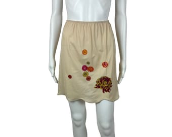 May include: A beige slip skirt with a scalloped hem and floral embroidery. The embroidery features a large pink and yellow flower with smaller orange and pink flowers surrounding it.