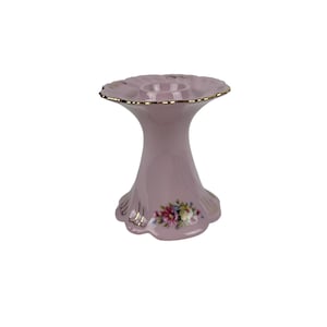 May include: A pink ceramic candle holder with a floral design and gold accents. The holder has a wide base and a narrow top with a circular opening for a candle.