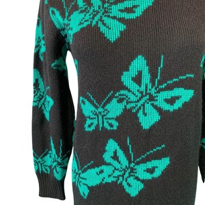 May include: A dark gray sweater dress featuring a repeating pattern of large, teal butterflies. The dress has long sleeves and a crew neck. The butterfly design is a striking contrast against the dark background, creating a bold visual statement.