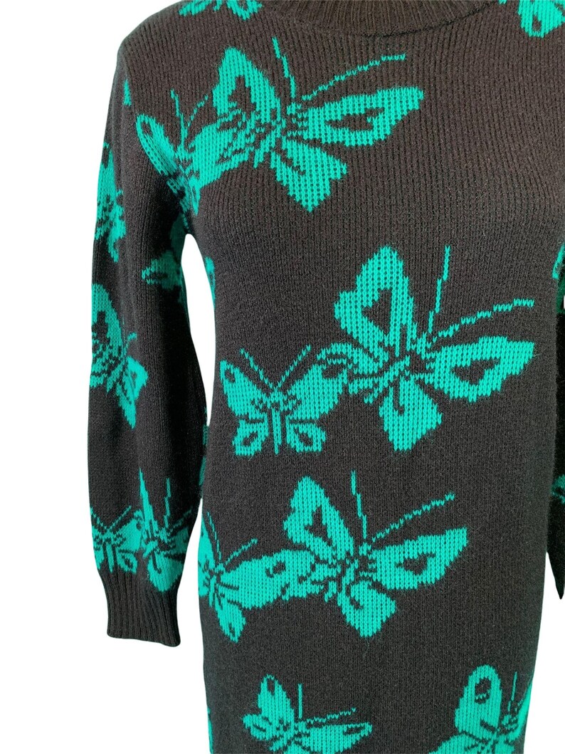 May include: A dark gray sweater dress featuring a repeating pattern of large, teal butterflies. The dress has long sleeves and a crew neck. The butterfly design is a striking contrast against the dark background, creating a bold visual statement.