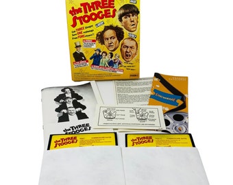 May include: A vintage-style box set for the Commodore 64 video game "The Three Stooges". The box set includes two game cartridges, a product catalog, and instruction manuals. The front of the box features a photo of the Three Stooges, Moe, Larry, and Curly.