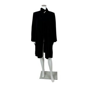 Puede incluir: Un abrigo largo de terciopelo negro con cuello alto y cierre decorativo. El abrigo se exhibe en un maniquí sobre un fondo blanco. El diseño del abrigo es sencillo y elegante.