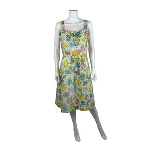 Könnte beinhalten: Ein Kleid mit Blumenmuster auf einer Schaufensterpuppe. Das Kleid hat einen weißen Hintergrund mit einem bunten Blumenmuster in Gelb-, Rosa-, Blau- und Grüntönen. Es hat einen quadratischen Ausschnitt, breite Träger und eine Bindetaille. Das Kleid reicht bis zur Mitte der Wade.