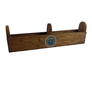 Könnte beinhalten: Ein hölzernes Wandregal mit rechteckigem Design und drei abgerundeten Griffen. Das Regal besteht aus hellbraunem Holz und hat einen runden Aufkleber mit dem Buchstaben "M" in einem blau-weißen Design.