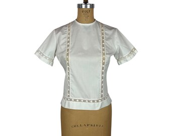 May include: A white short-sleeved blouse with decorative lace trim down the front and around the sleeves. The blouse is displayed on a vintage dress form. The dress form is tan and has the words "COLLAPSIBLE MODEL 1961" printed on it.