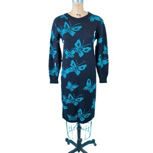 May include: A navy blue sweater dress with a repeating turquoise butterfly pattern. The long-sleeved dress is displayed on a vintage dress form. The dress has a crew neck and a knee-length hemline. The dress is made of a knitted material.