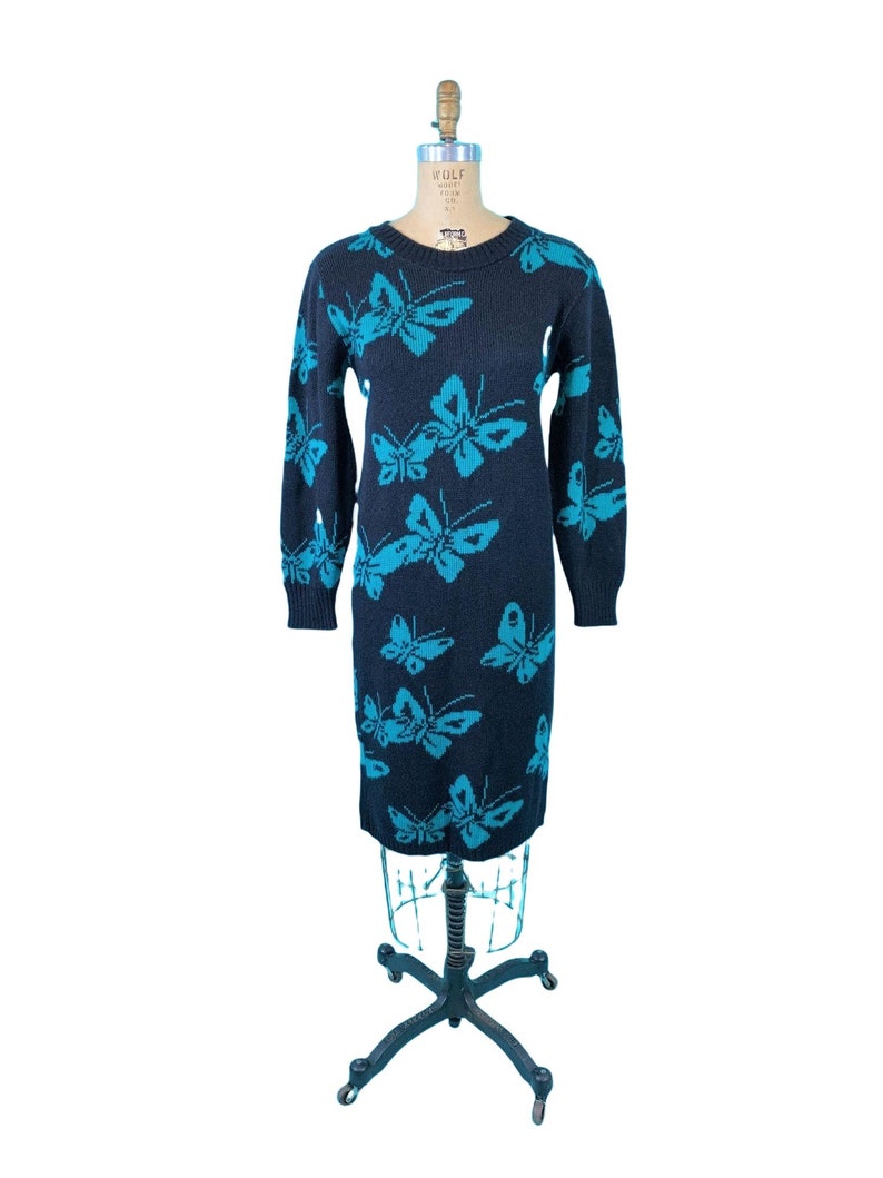 May include: A navy blue sweater dress with a repeating turquoise butterfly pattern. The long-sleeved dress is displayed on a vintage dress form. The dress has a crew neck and a knee-length hemline. The dress is made of a knitted material.