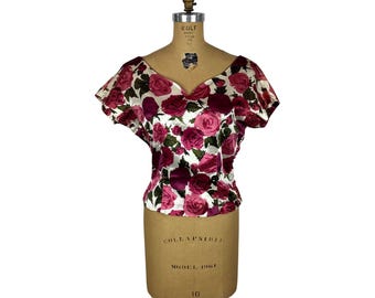 May include: A vintage floral print top with a sweetheart neckline. The top features a white background with a pattern of pink and burgundy roses and green leaves. The garment is displayed on a vintage dress form.