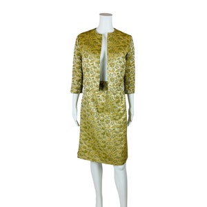 May include: A vintage two-piece suit in a gold and green floral brocade fabric. The jacket has a collarless neckline and three-quarter sleeves. The skirt is knee-length and has a straight silhouette.