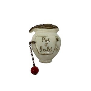 May include: White ceramic pot of gold trinket with a gold chain and red bead. The pot has a lid with gold coins and the words "Pot o' Gold" written on the front.