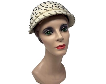 May include: A cream-colored hat with a rounded crown and a small brim. The hat is adorned with small, dark-colored embellishments and a decorative trim. The hat is displayed on a mannequin head with dark eye makeup and red lipstick.