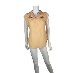May include: A peach-coloured, short-sleeved blouse with a collared neckline. The blouse has embroidered floral details in red, orange, and green near the neckline. The garment is displayed on a mannequin.