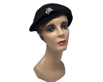 May include: A black beret with a decorative rhinestone brooch. The beret is displayed on a mannequin head with a neutral skin tone, red lipstick, and green eyes. The brooch is a small, ornate design.