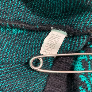 May include: Close-up of a black and teal knit fabric with a safety pin. A white tag with care instructions is attached. The tag reads "100% Acrylic" and "Made in U.S.A."