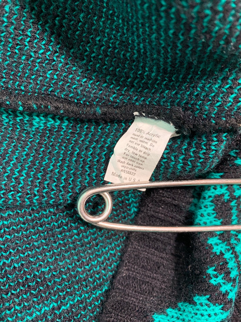 May include: Close-up of a black and teal knit fabric with a safety pin. A white tag with care instructions is attached. The tag reads "100% Acrylic" and "Made in U.S.A."