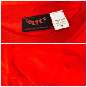 May include: Red t-shirt with a black tag that reads "COLTEX" and "THE WALT DISNEY COMPANY". The tag also includes care instructions and size "M".