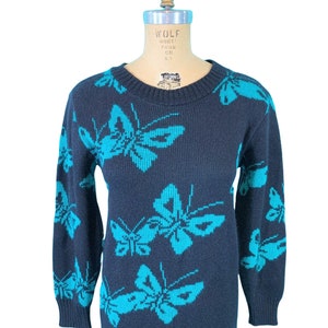 May include: Navy blue sweater dress featuring a repeating pattern of turquoise butterflies. The long-sleeved dress has a crew neck and is displayed on a mannequin. The butterfly design is visible on the sleeves and body of the dress.