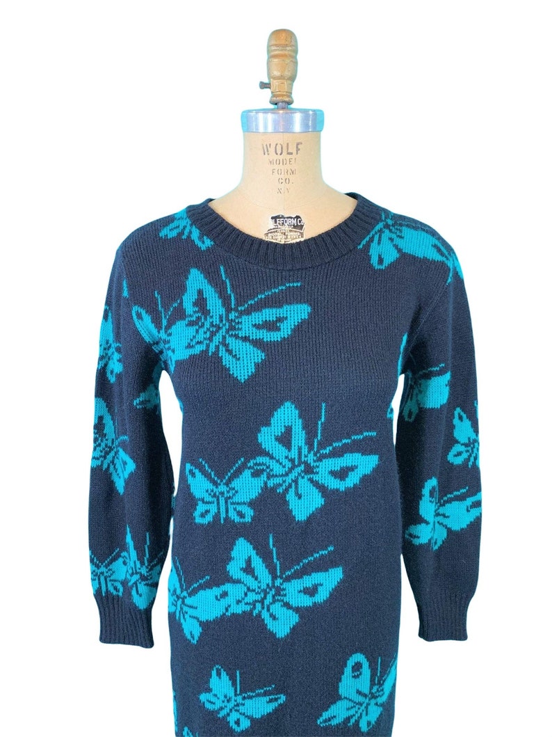 May include: Navy blue sweater dress featuring a repeating pattern of turquoise butterflies. The long-sleeved dress has a crew neck and is displayed on a mannequin. The butterfly design is visible on the sleeves and body of the dress.