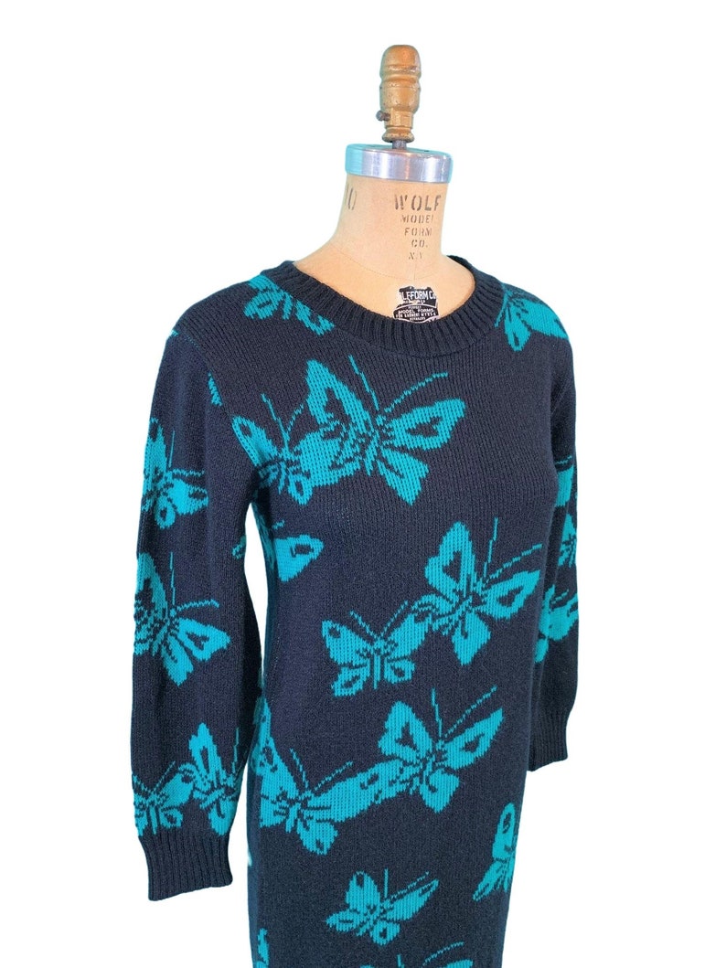 May include: A navy blue knit dress featuring a repeating pattern of turquoise butterflies. The long-sleeved dress has a crew neck and a knee-length hemline. The dress is displayed on a vintage dress form.